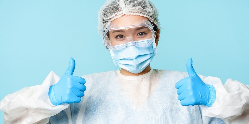 Portrait of smiling asian nurse or doctor, wearing personal protective equipment, showing thumbs up, recommending, standing over blue background