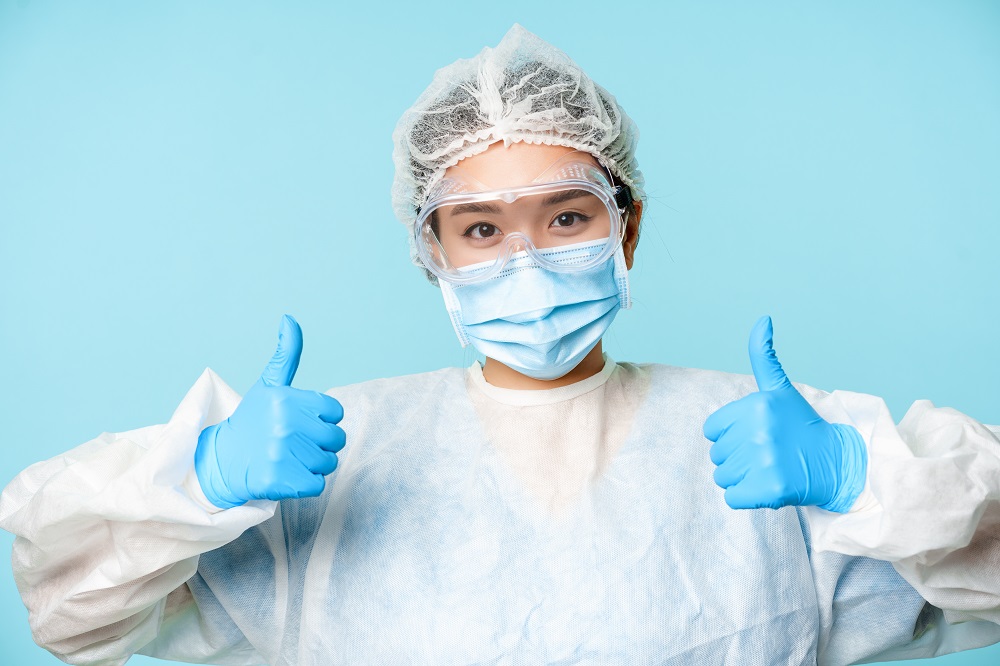 Portrait of smiling asian nurse or doctor, wearing personal protective equipment, showing thumbs up, recommending, standing over blue background
